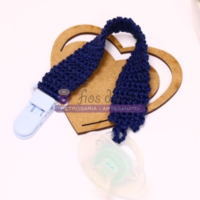 Fita em crochê azul escura com clip branco e chupeta transparente sobre suporte de madeira em forma de coração