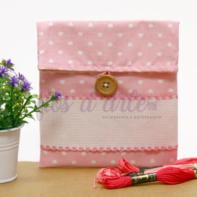 Bolsa rosa com corações brancos, botão de madeira e renda rosa junto a dois novelos de linha vermelha e vaso branco com flores roxas