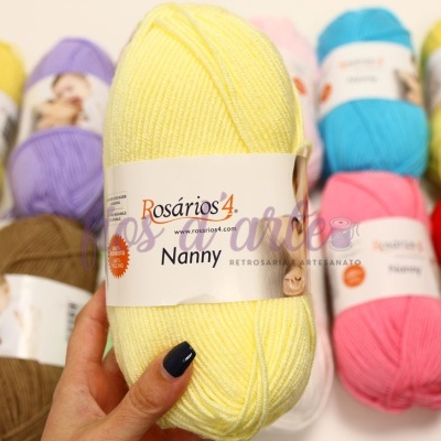 Novelo de lã amarelo da marca Rosários 4 modelo Nanny segurado por mão com unhas azuis, fundo com mais novelos coloridos.