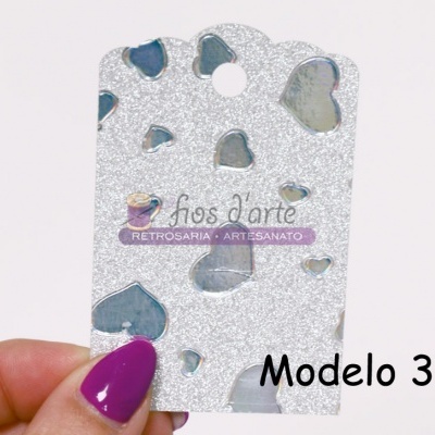 Cartão brilhante prateado com recortes de corações e texto MODELO 3, segurado por mão com unhas roxas