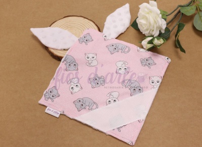 Manta bebé rosa com padrão de gatos e orelhas brancas felpudas junto a flores brancas e base de madeira