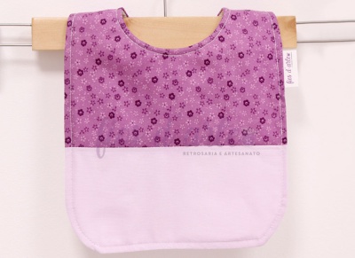 Babete lilás com padrão de flores e parte inferior branca, pendurado numa roupa de madeira.