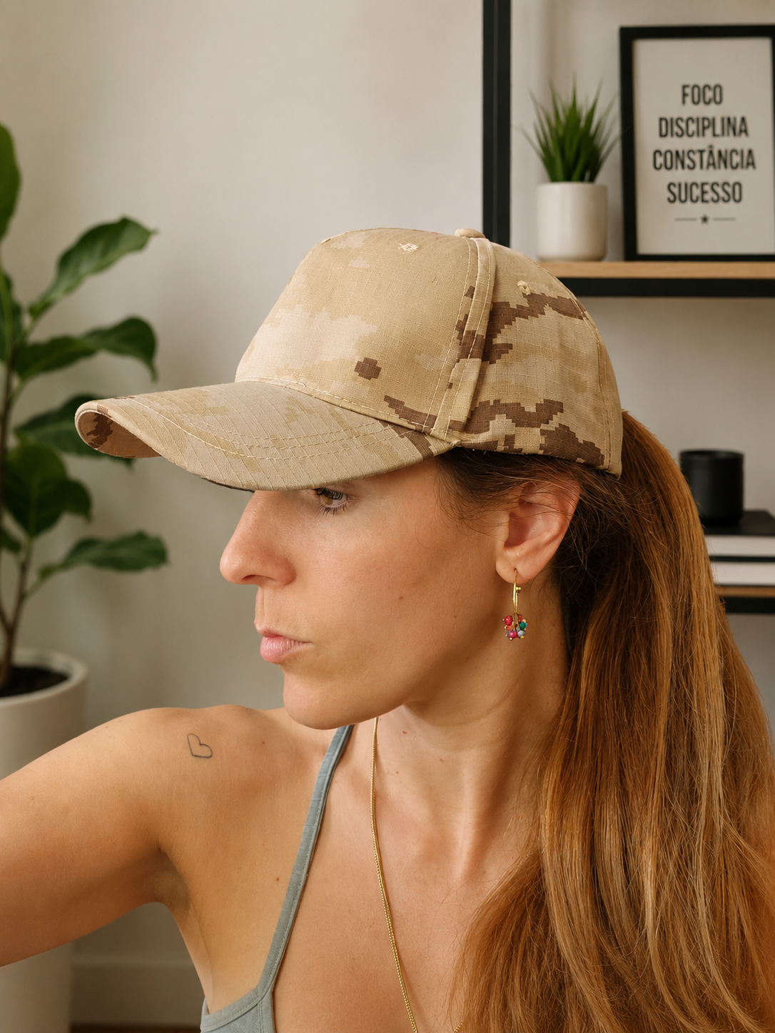 Boné camuflado bege usado por mulher com brincos coloridos e cabelo preso em ambiente interior com planta e quadro.