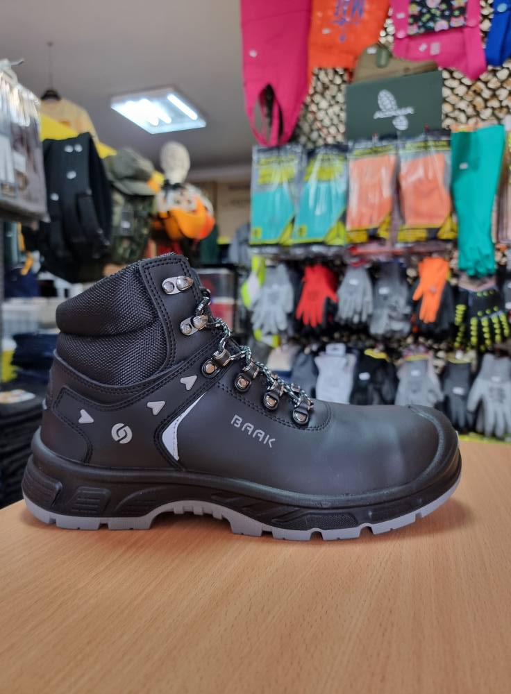 Bota de segurança preta com sola cinza sobre mesa de madeira com luvas penduradas no fundo