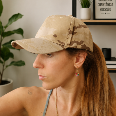 Boné camuflado bege usado por mulher com brincos coloridos e cabelo preso em ambiente interior com planta e quadro.