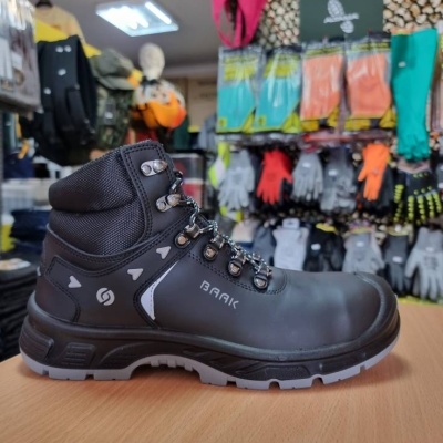 Bota de segurança preta com sola cinza sobre mesa de madeira com luvas penduradas no fundo