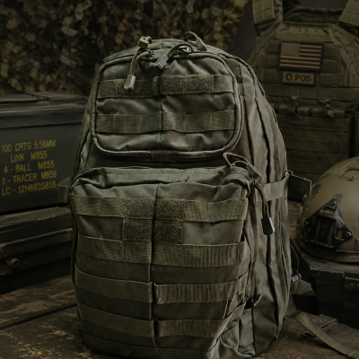 Mochila militar verde-oliva tática com vários bolsos e tiras, sobre caixa de madeira em ambiente militar