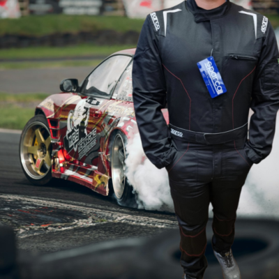 Fato de corrida preto com etiquetas SPARCO, pessoa em pé e carro de corrida a derrapar ao fundo