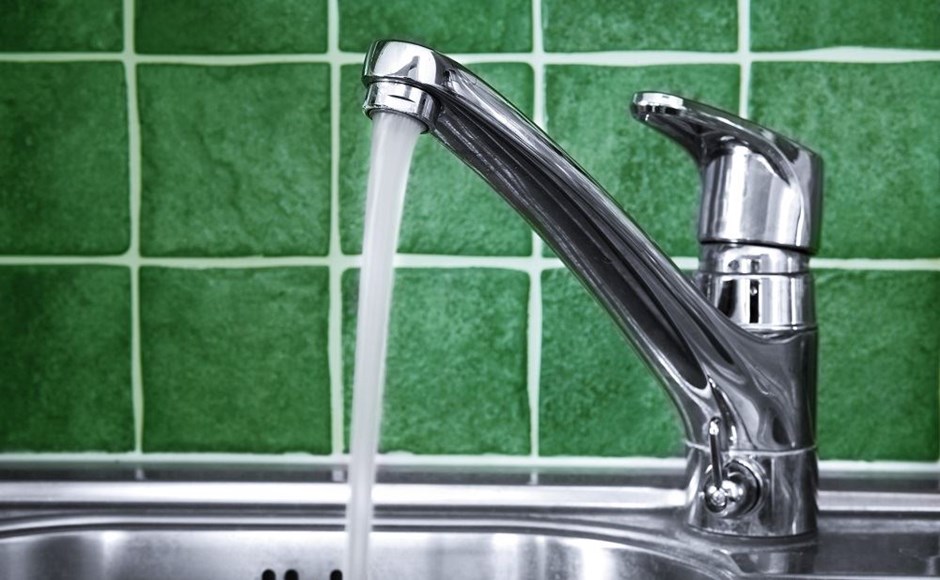 Torneira de cozinha cromada com água a correr e azulejos verdes ao fundo