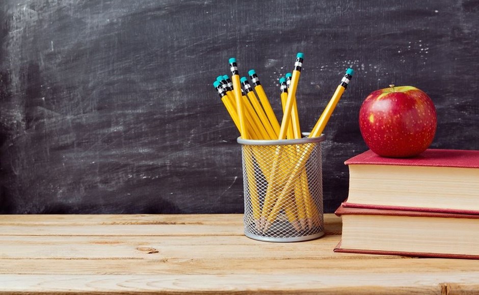 Lápis em suporte metálico, maçã vermelha e livros sobre mesa com quadro negro ao fundo