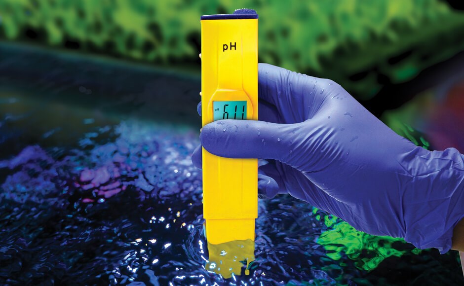 Medidor de pH digital amarelo na mão com luva roxa sobre líquido azul esverdeado