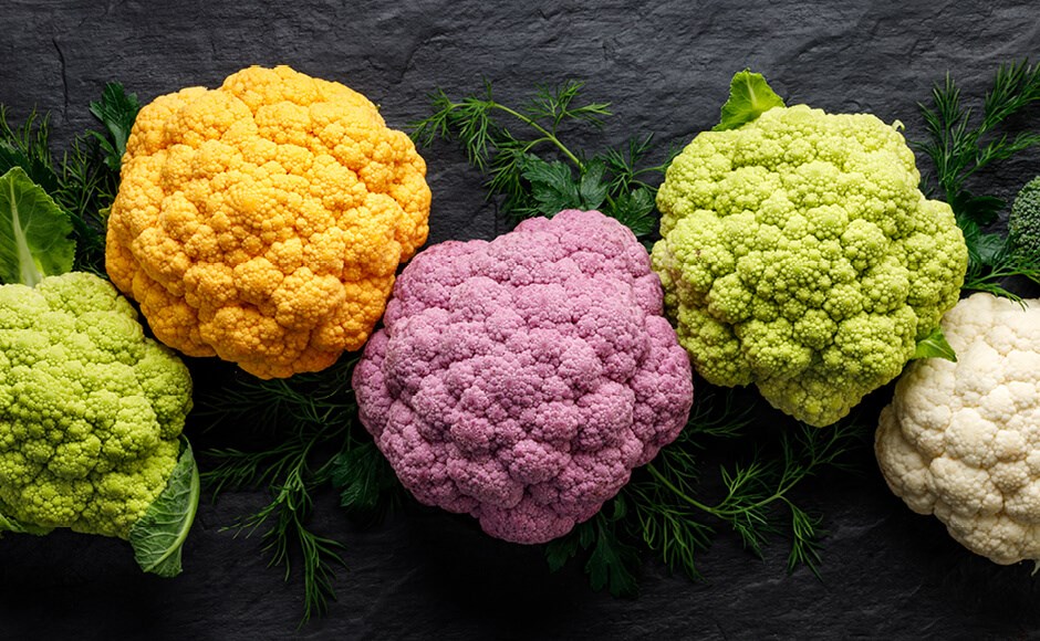 Cinco couves-flor coloridas sobre fundo escuro com ervas verdes