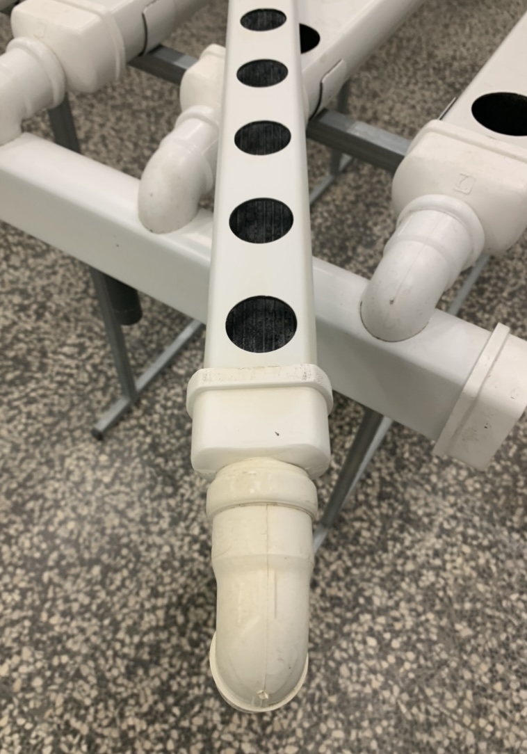 Joelho para Topos de Descarga - 2 mão Estrutura tubular branca em PVC com orifícios circulares e suportes metálicos