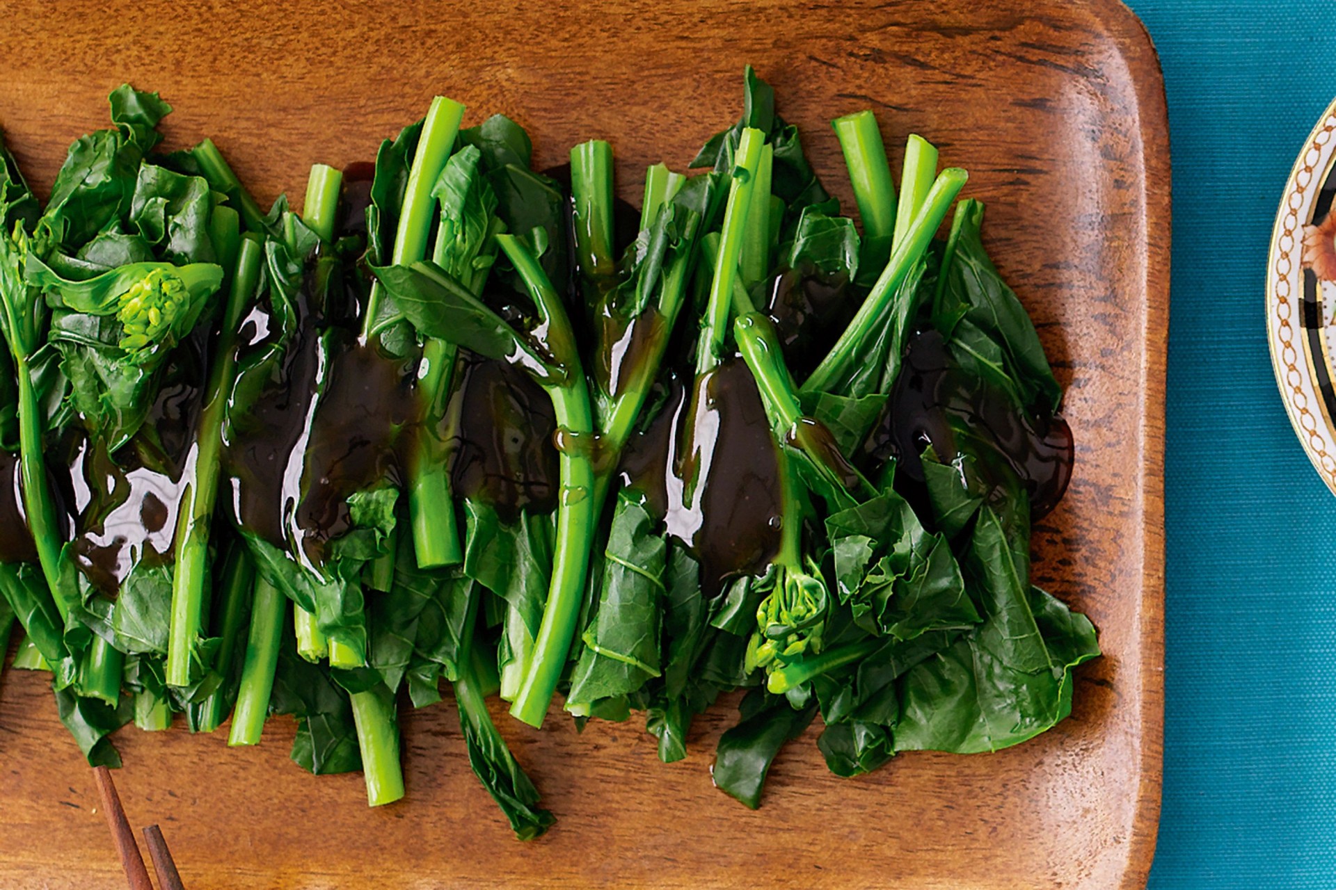 Legumes verdes com molho escuro sobre prato de madeira, pauzinhos e prato ao lado em superfície azul