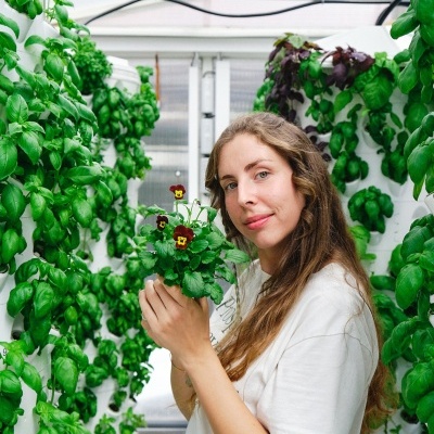 Mulher segurando planta com flores roxas entre plantas verdes em estufa