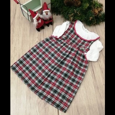 Vestido infantil xadrez vermelho verde branco preto com blusa branca manga comprida, chão madeira, bonecos Papai Noel e arranjo ramos verdes com pinhas
