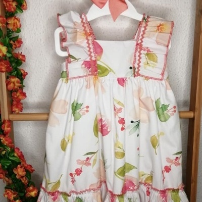 Vestido infantil branco com padrão floral colorido e babados pendurado num cabide branco com laço rosa.