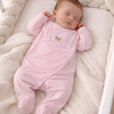 Bebé a dormir em saco de dormir cor-de-rosa com bordado de cavalo de madeira e laços num berço com proteção trançada creme.