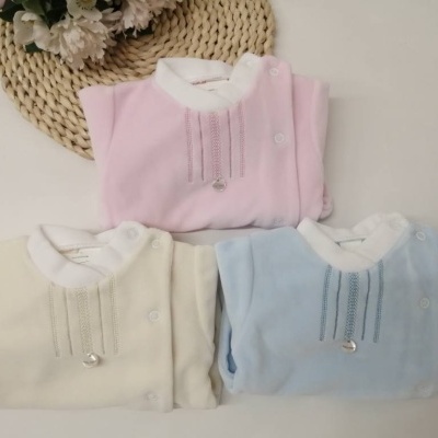 Três robes de bebé rosa, creme e azul claro com bordados e enfeite prateado, dispostos sobre uma superfície branca com tapete de palha e flores brancas.