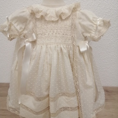 Vestido de bebé branco com padrão de pompons, renda e fitas de cetim num manequim.