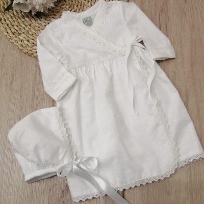 Vestido branco de bebé com renda e laço sobre mesa de madeira com flores e tapete de palha.