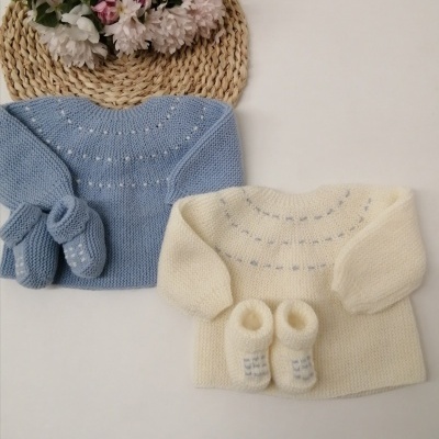 Conjunto de roupa de bebé em malha azul e creme com padrão de pontos, apresentado sobre fundo branco com tapete de palha e flores.