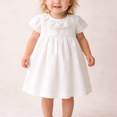 Criança com vestido branco e sapatos brancos em fundo neutro.