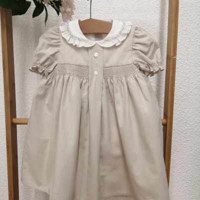 Vestido infantil bege com gola branca bordada pendurado em cabide numa estrutura de bambu