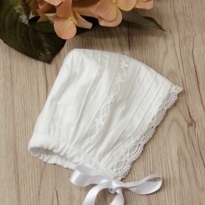 Gorro de bebé branco com renda e fita de cetim sobre madeira clara e flores artificiais