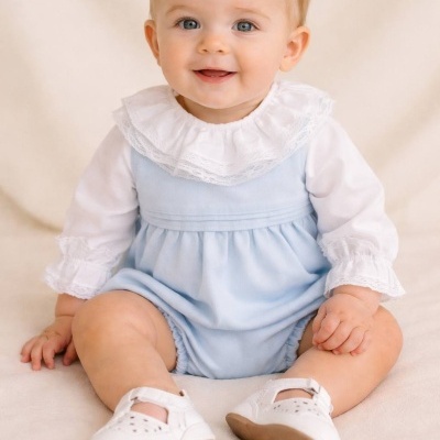 Bebé vestido de azul claro com gola branca em renda e sapatos brancos sentada.