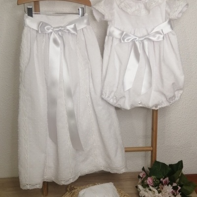 Conjunto infantil branco com vestido, saia, fita prateada e touca, com detalhes em renda e bolinhas, em cenário de madeira e flores.