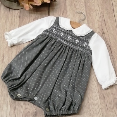 Roupa infantil com vestido cinza de pontos brancos sobre blusa branca, pendurada em cabide sobre chão de madeira
