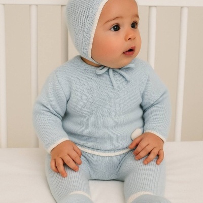 Bebé vestido com conjunto de malha azul claro sentado numa cama branca.