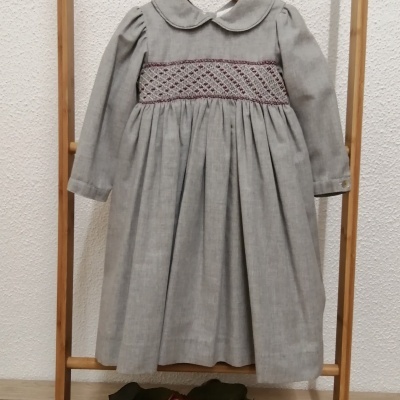 Vestido infantil cinza com bordo roxo pendurado em estrutura de madeira com flores no chão