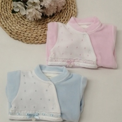 Dois fatos de bebé rosa e azul com pequenos detalhes e laços, sobre superfície branca, ao lado de flores num tabuleiro de palha.