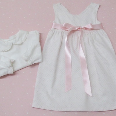 Conjunto branco de vestido com fita rosa e camisola delicada para bebé sobre fundo rosa claro.