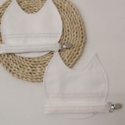 Dois babetes brancos de tecido com cintos ajustáveis e prendedores metálicos, sobre fundo branco e base de fibras naturais entrelaçadas