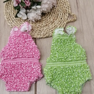 Dois vestidos de bebé rosa e verde com textura felpuda sobre suporte de rattan e superfície de madeira clara com flores artificiais.