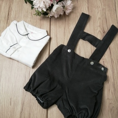 Conjunto infantil com camisa branca e calções pretos com alças sobre chão de madeira clara e flores artificiais.