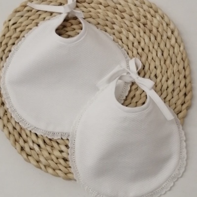 Dois babetes brancos para bebé com renda e fita sobre suporte trançado
