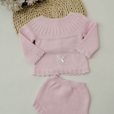 Conjunto de bebé cor-de-rosa em malha com camisola e cueiro, flores ao fundo.