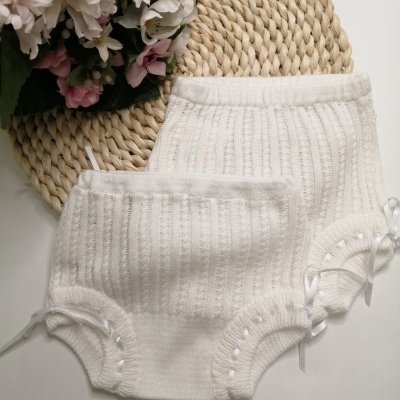 Dois calções de malha branca para bebé com padrão canelado e laços laterais em fundo branco com base redonda de fibra natural e flores cor-de-rosa.