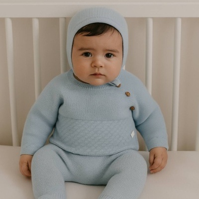 Bebé com roupa de malha azul clara sentado numa cama branca.