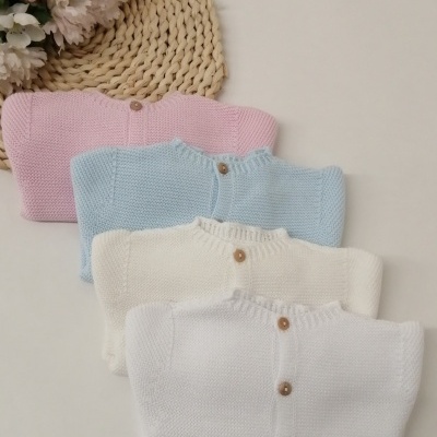 Cardigans de malha para bebé em rosa, azul, creme e branco com botões de madeira, junto a flores e naperon