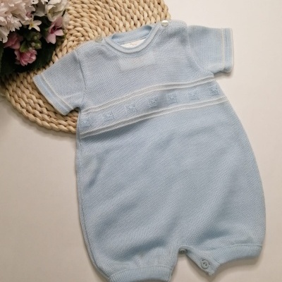 Macacão curto azul claro de bebé em malha texturizada com botões, sobre tapete de palha com flores ao lado.
