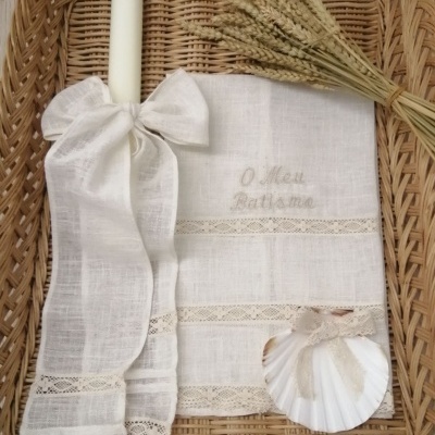 Conjunto branco de batismo com vela, toalha bordada, concha e ramo de trigo numa cesta de verga