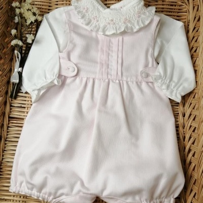 Macacão de bebé rosa claro com mangas brancas e gola rendada, tecido macio, colocado sobre base de rattan.