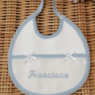 Babete branco com borda azul clara e nome 'Francisco' bordado, sobre rattan com flores ao fundo