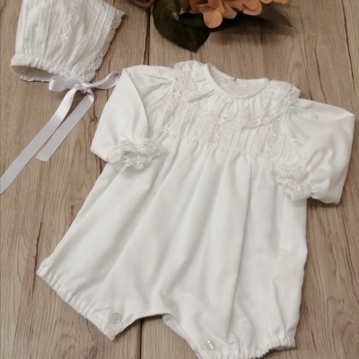 Macacão branco para bebé com detalhes em renda e chapéu branco combinando sobre fundo de madeira.
