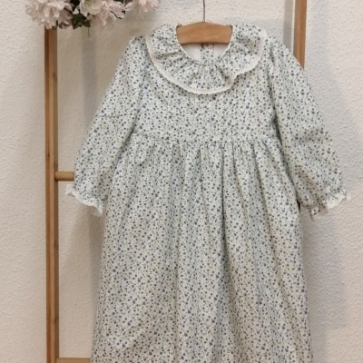 Vestido infantil branco com padrão floral, gola com folhos, mangas compridas, pendurado em estrutura de madeira com flores decorativas
